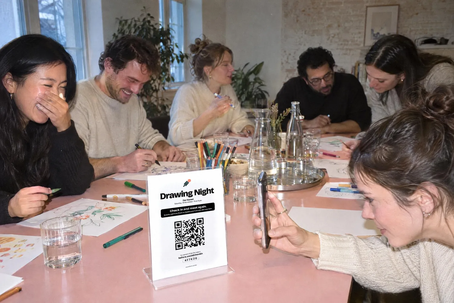A drawing night in a bright room at a long pink table: guests draw and laugh, in front the card with the QR code stands in a holder and a guest scans it with her phone