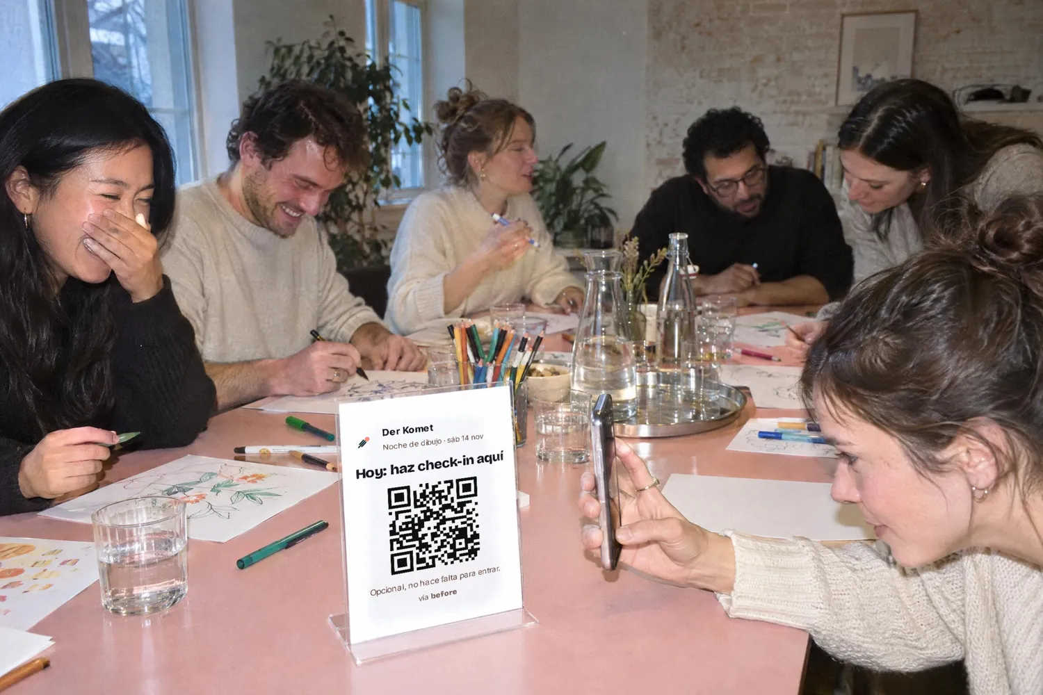 Una noche de dibujo en una sala luminosa, en una mesa larga de color rosa: los invitados dibujan y ríen; delante, la tarjeta con el código QR está en un soporte y una invitada la escanea con su teléfono