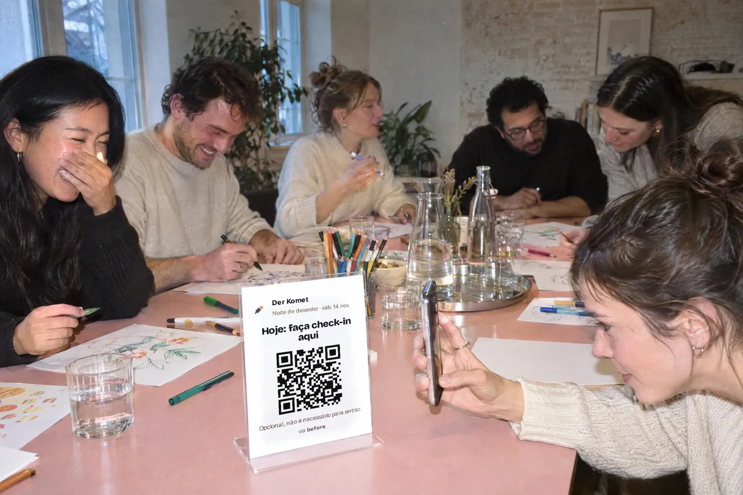Uma noite de desenho numa sala clara, numa mesa comprida cor-de-rosa: convidados desenham e riem, na frente o cartão com o QR code está num suporte e uma convidada o escaneia com o celular