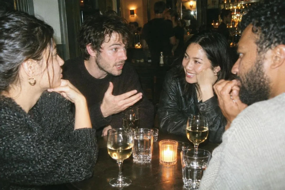 Quatre invités discutent autour d'une petite table ronde de bar, l'un explique avec la main ouverte, une autre écoute, le menton dans la main