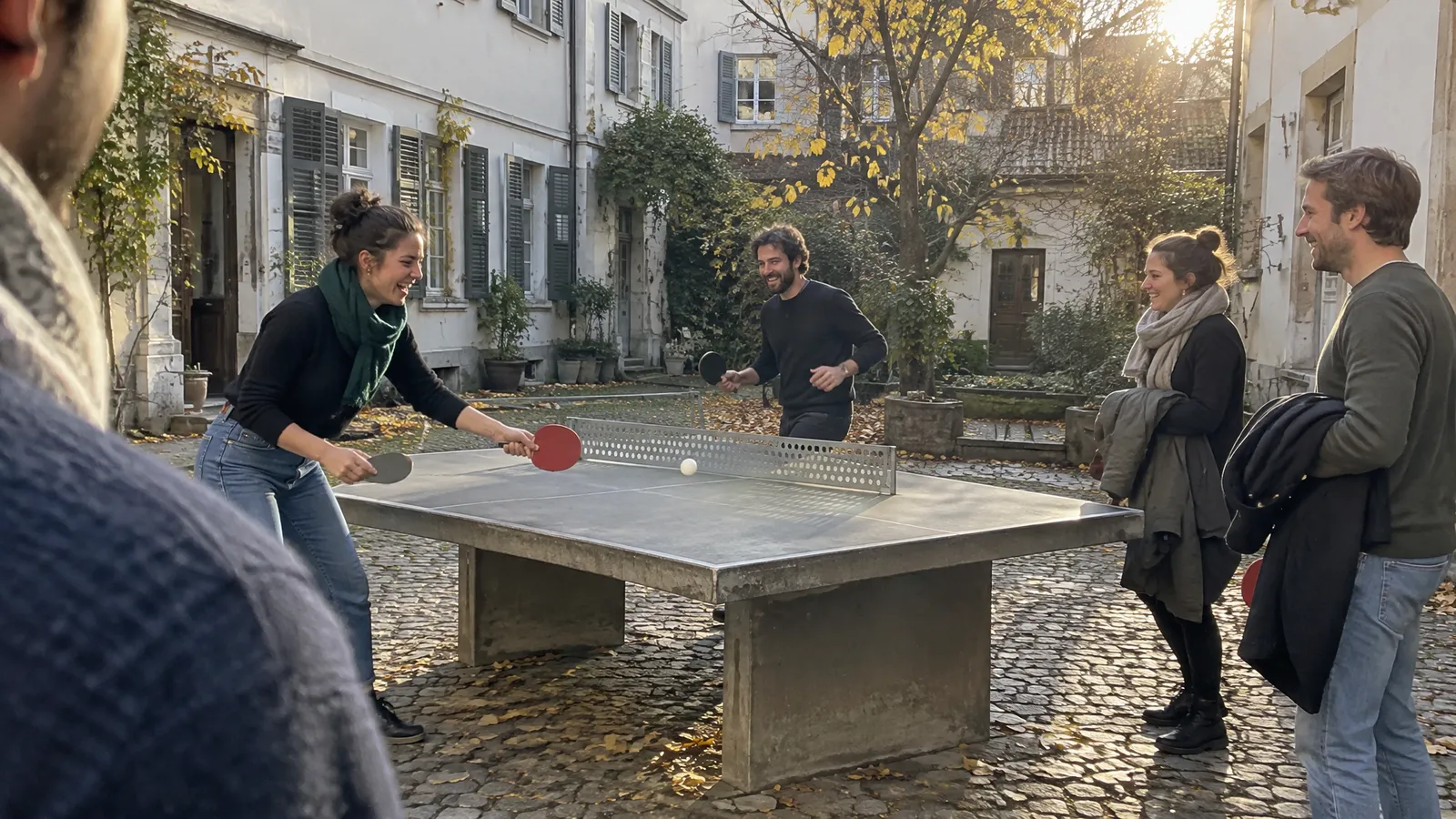 People playing round-the-table ping pong at a concrete table in a Basel courtyard on a late autumn afternoon; one laughs as she reaches for the ball, one runs round the table, two wait their turn with jackets over their arms