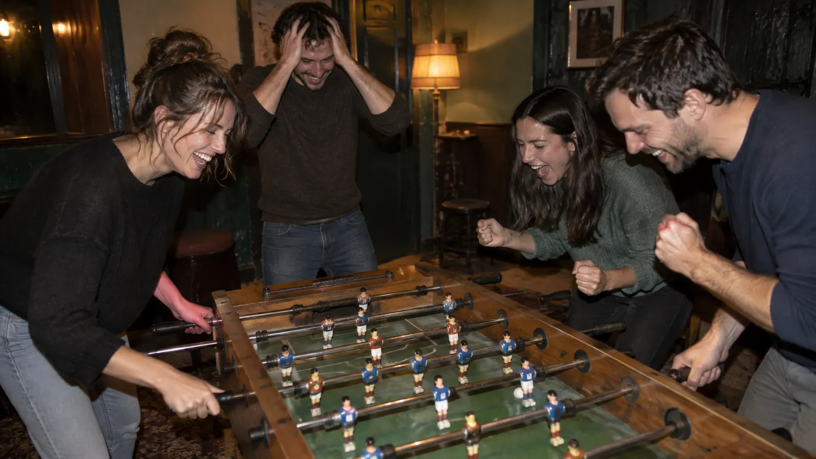 Four friends in their late twenties and thirties around a well-worn table football game in the back of a small bar on an autumn evening; one woman laughs as she spins her rod, a man clutches his head with both hands, two lean over the edge cheering, warm lamp light