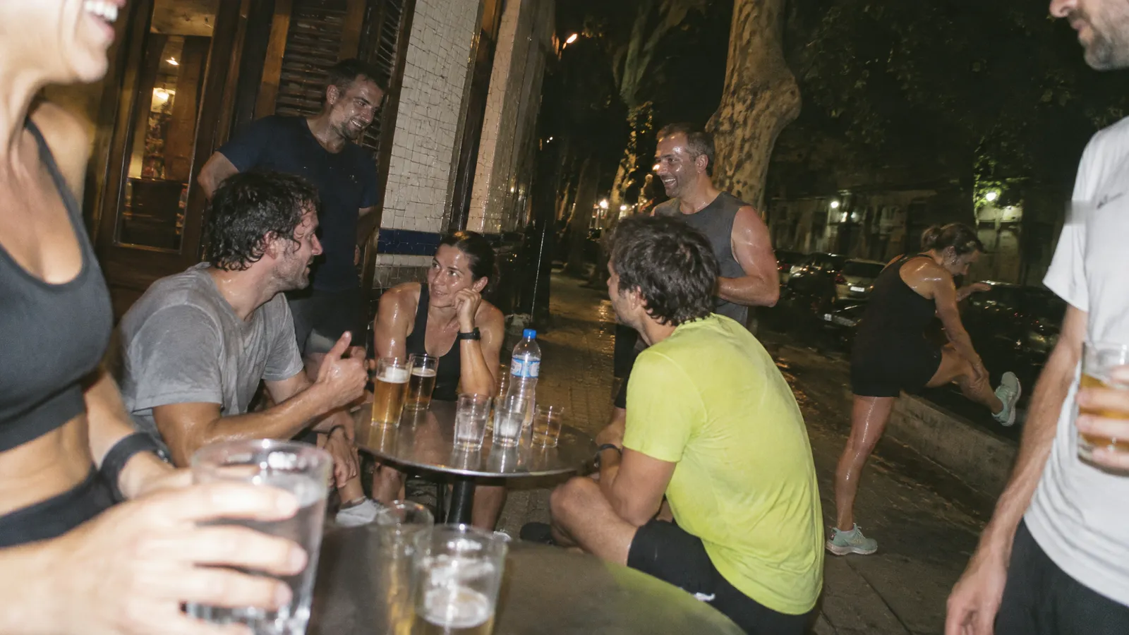 Corredores todavía transpirados alrededor de dos mesitas en la vereda de un bar de esquina en Buenos Aires, de noche, con vasos de cerveza y de agua; una corredora estira la pierna en el cordón y arriba se ven los plátanos de la calle