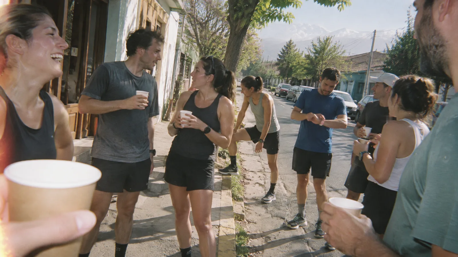 Corredores con vasos de papel con café en la vereda frente a una cafetería de Santiago, una mañana de sol: dos conversan, una estira la pierna en la cuneta, otro mira su reloj y al fondo se ve la cordillera nevada