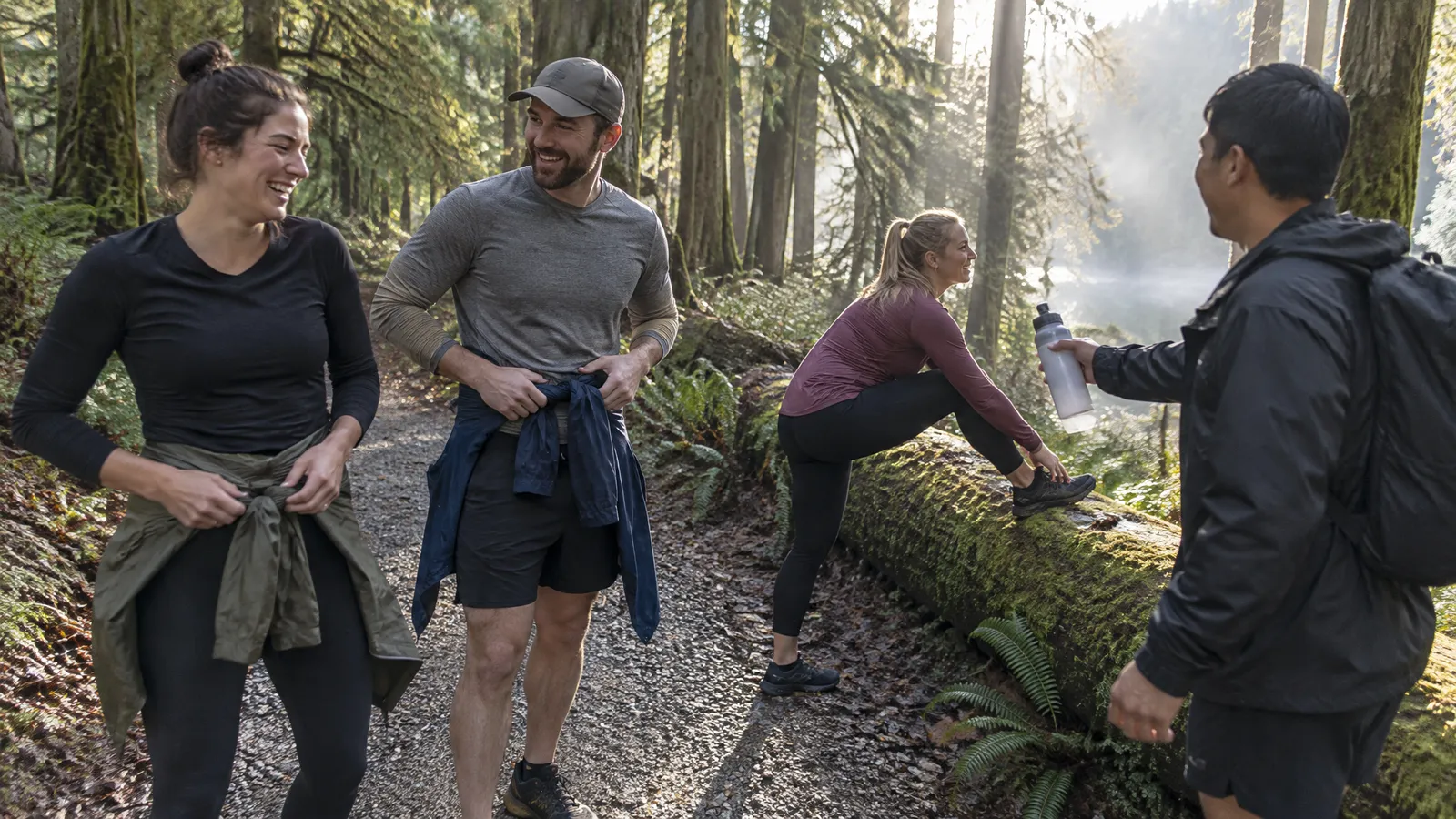 Runners in their late twenties and thirties catching their breath at the end of a gravel trail among tall cedars and ferns on a bright, misty fall morning; two laugh as they tie damp rain jackets around their waists, one stretches her calf against a mossy fallen log, one hands a water bottle along, sunlight slanting through the trees