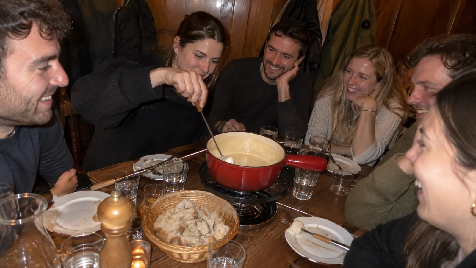 Six guests between their mid-twenties and late thirties sit around a cheese fondue pot in the wood-panelled back room of a small Zurich restaurant on an autumn evening; one woman fishes for a lost piece of bread with her long fork while the others laugh, with water glasses and a bread basket on the table and jackets hanging on hooks on the panelled wall